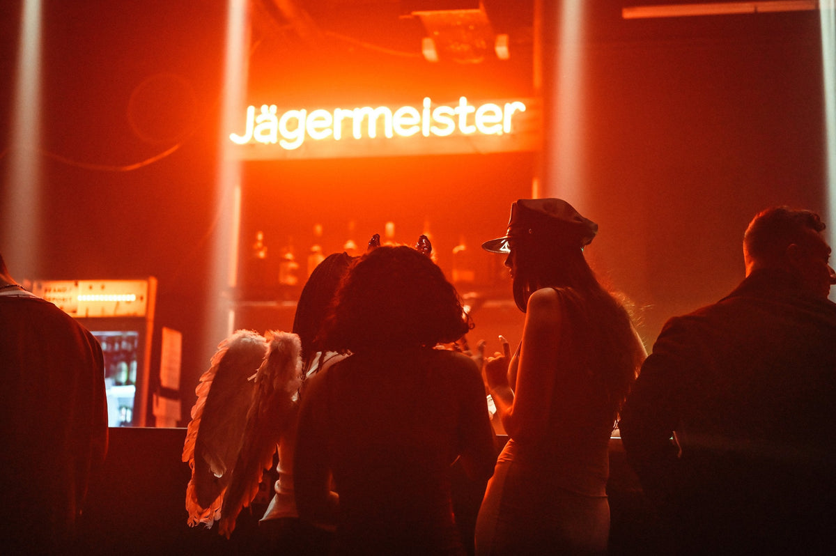 A dimly lit nightclub or bar scene with several silhouetted people facing a glowing neon sign that reads 'Jägermeister'. The scene is dominated by warm orange-red lighting, with one person wearing angel wings and another wearing a hat.