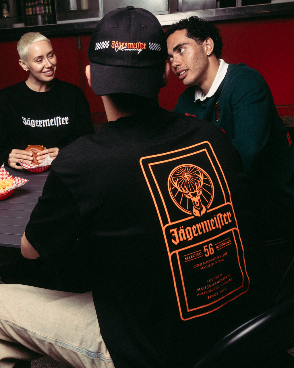 Four people gathered around a table at a food stall, with the central focus on a person wearing a black Jägermeister t-shirt with a large orange graphic on the back, khaki pants, and a black cap. 