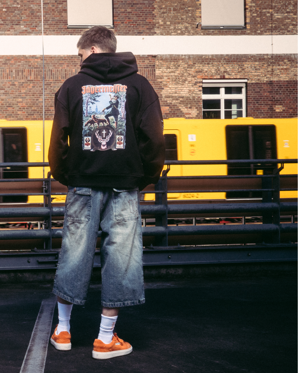 A model wearing streetwear apparel, including a black hoodie with the Jägermeister tradition graphic back print, standing on a dark paved surface in front of a brick building and a yellow train.
