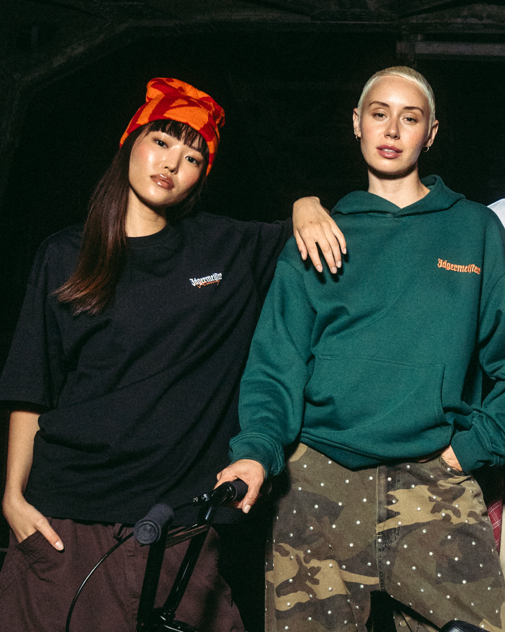 Two young women posing with a black BMX bike in front of a dark industrial bridge at night. They are dressed in Jägermeister streetwear, including a green hoodie, white t-shirt, black vest, and cargo pants.