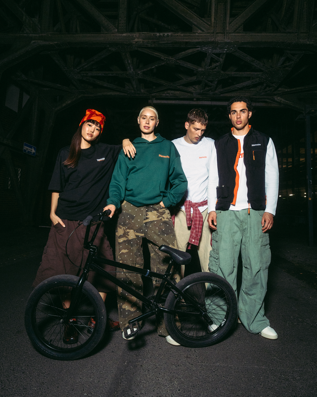 Four young men posing with a black BMX bike in a dark, industrial bridge setting at night. They are dressed in Jägermeister streetwear outfits, including a green hoodie, white t-shirt, black vest, and cargo pants.