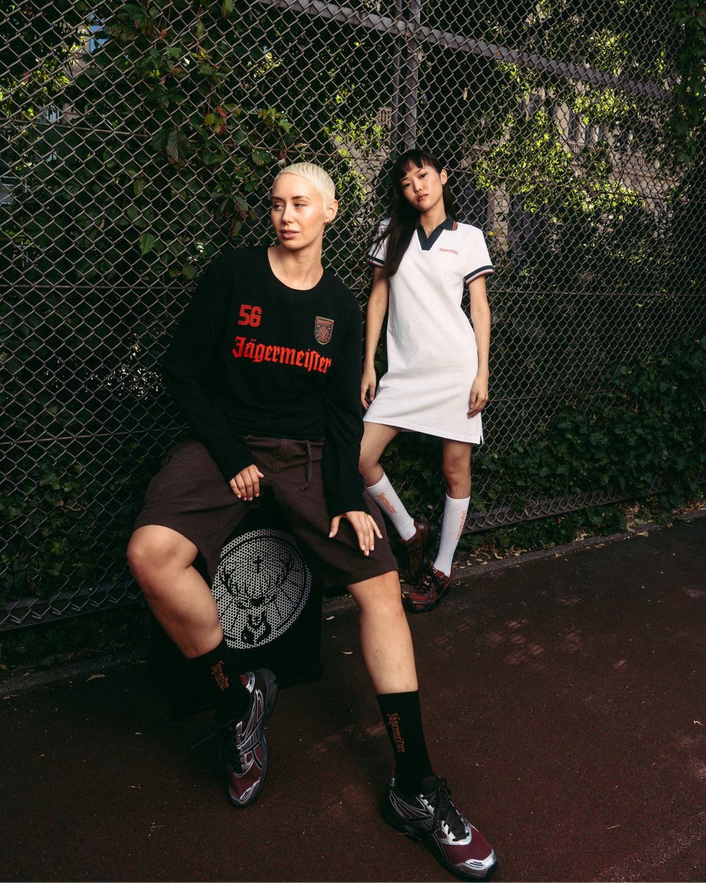 Two models posing in front of a chain-link fence. The model on the left wears a black Jägermeister long-sleeve, brown shorts, and black socks.