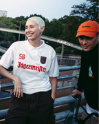 Cuatro personas de pie al aire libre sobre un puente o pasarela, llevando streetwear de Jägermeister. Una lleva una chaqueta naranja brillante con cremallera, otra lleva una camiseta tipo jersey verde oscuro de Jagermeister con una falda blanca, una tercera lleva una camiseta tipo jersey blanca de Jagermeister con pantalones cortos oscuros, y otra lleva una sudadera negra de Jägermeister y una gorra naranja.