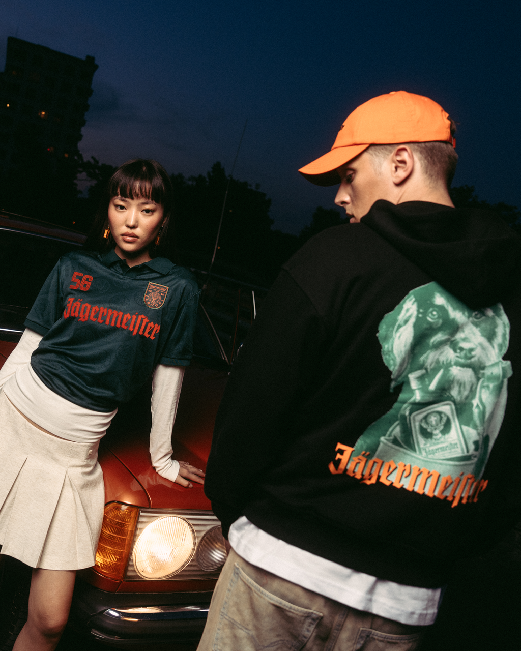 Dos modelos posan delante de un coche rojo vintage al atardecer. Una modelo lleva una camiseta tipo jersey verde oscuro de 'Jägermeister' con una falda blanca plisada, y la otra lleva una sudadera con capucha negra de Jägermeister Waldi y una gorra naranja.