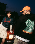 Dos modelos posan delante de un coche rojo vintage al atardecer. Una modelo lleva una camiseta tipo jersey verde oscuro de 'Jägermeister' con una falda blanca plisada, y la otra lleva una sudadera con capucha negra de Jägermeister Waldi y una gorra naranja.