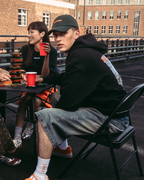 Cuatro personas sentadas alrededor de una pequeña mesa en una azotea o puente al aire libre, sosteniendo vasos rojos. Llevan streetwear informal de Jägermeister, incluida una sudadera naranja, una camiseta blanca, una sudadera con capucha negra y pantalones estampados.