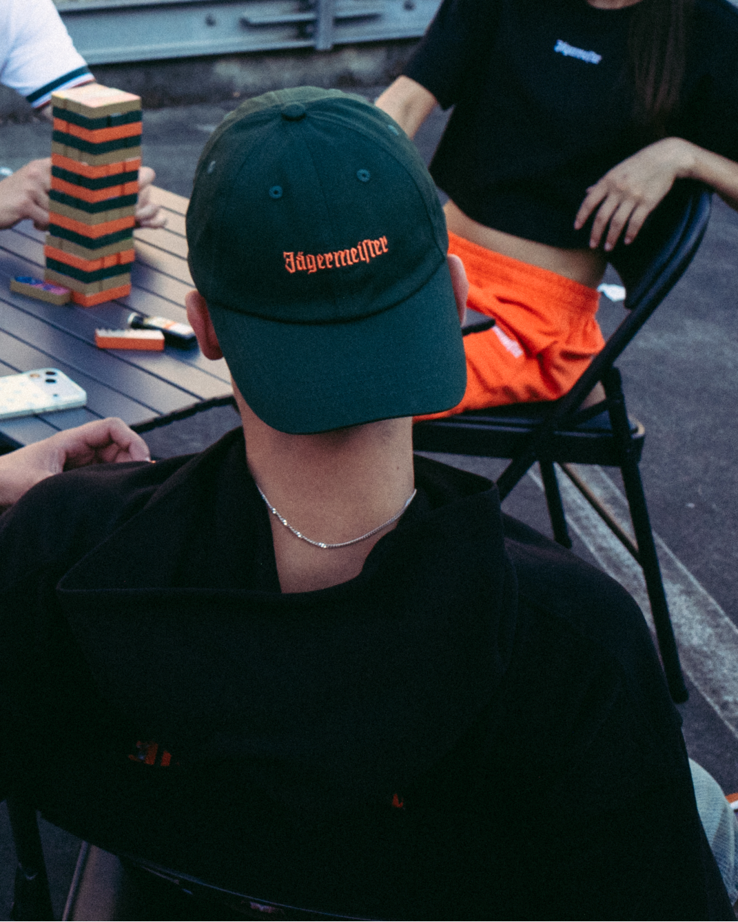 A smiling woman in the foreground wears a bright orange long-sleeve sweatshirt with '56 Jägermeister' printed in black on the front, holding a gold chain. Three people are seated at a table in the background on an outdoor rooftop or deck, with a brick building behind them.