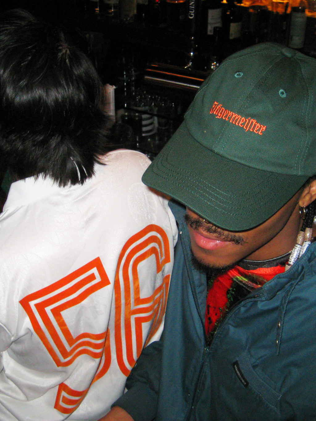 Dos personas sentadas juntas en un bar, una de ellas con una gorra de papá verde de Jägermeister.