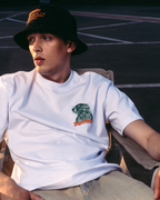 A man wearing a white graphic t-shirt with a Jägermeister Waldi logo, a black bucket hat, and beige jeans, sitting in a chair in an outdoor setting.