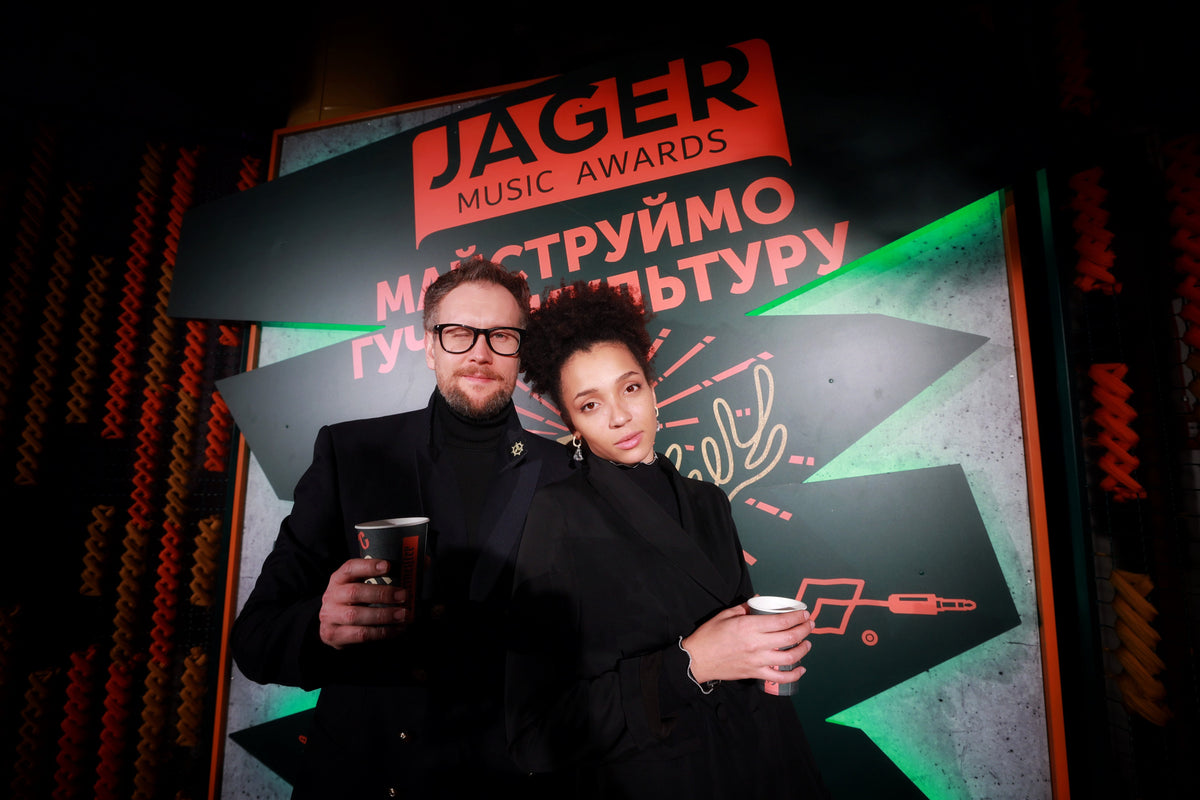 Two people standing in front of a large Jäger Music Awards backdrop at a night event, both wearing black and holding paper cups.