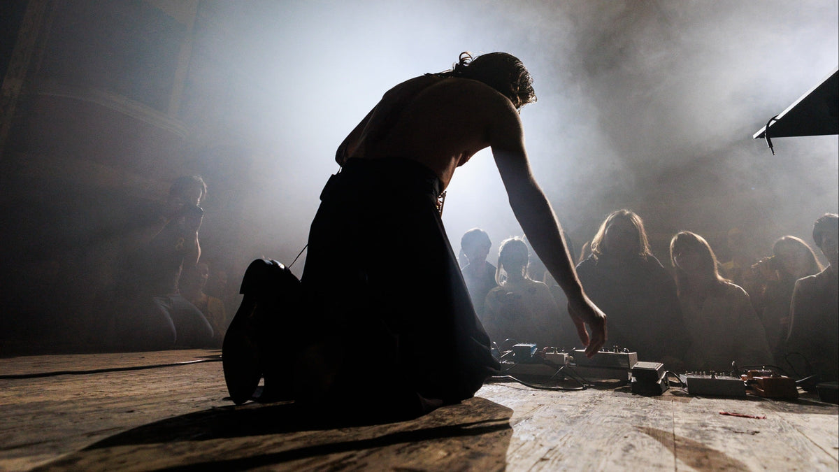 A shirtless person kneeling on a wooden floor in a smoky indoor setting, reaching toward small equipment in the foreground, with blurred figures in the background.