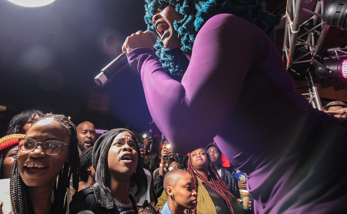 A young female with a purple dress is singing in front of a rather big crowd of people.