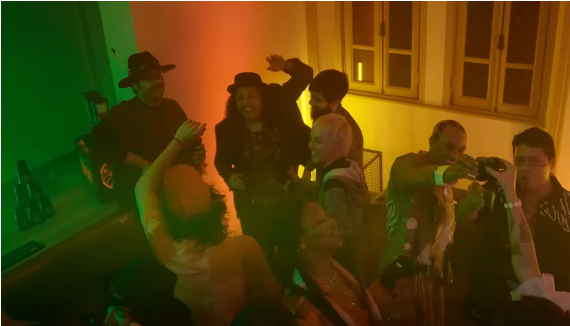 A group of people dancing and socializing in a dimly lit room with dramatic green and orange ambient lighting. Several individuals are visible from the back or side, wearing dark clothing, with one person in a black hat and another with a pink hairpiece.