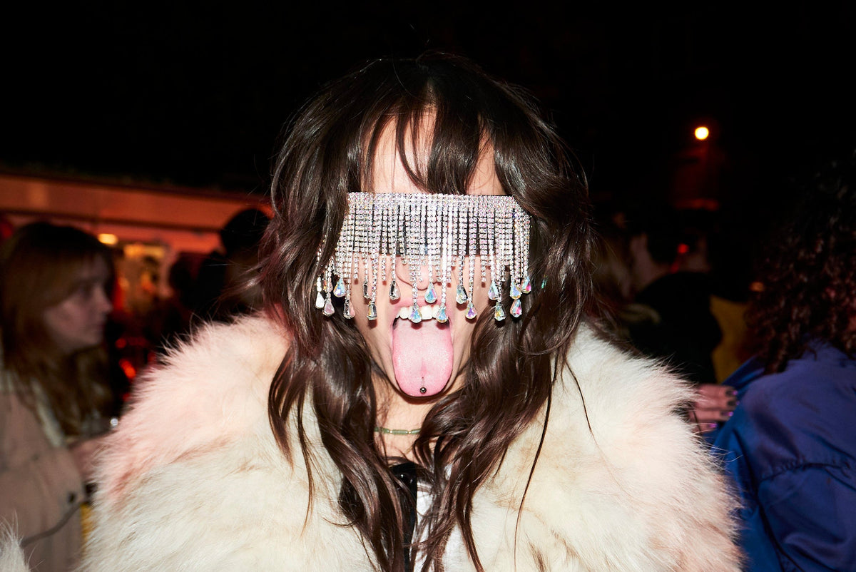 A person at a night event wearing a white fur coat and a silver rhinestone face mask with dangling charms, sticking their tongue out.