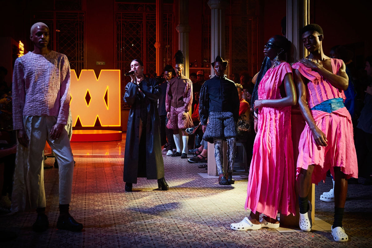 A fashion runway scene with multiple models walking in an indoor venue. In the foreground, a model wears a bright pink draped dress or tunic with a matching pink skirt, while another model beside them wears a pink patterned top and skirt with a blue sash.