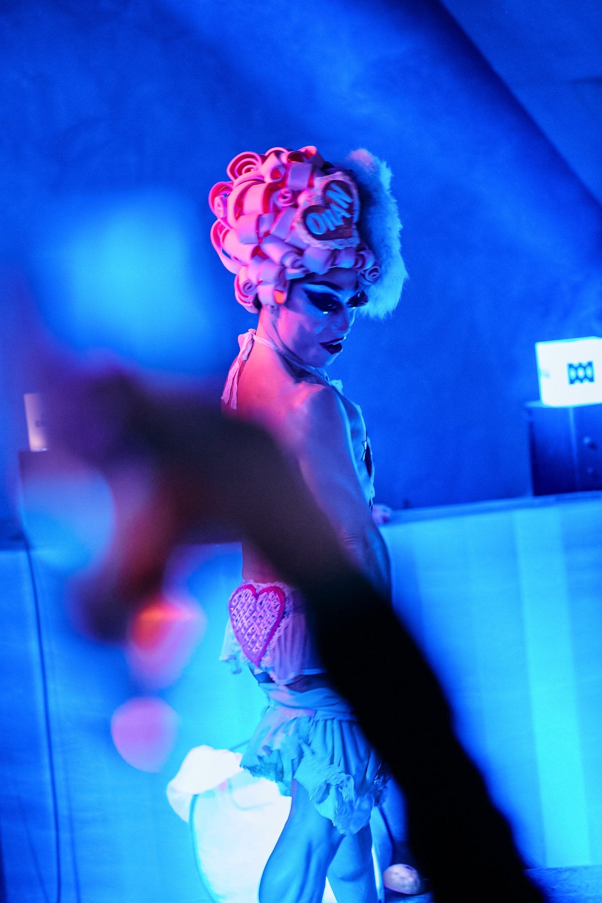 A person in a stylized costume with a large pink and white sculpted headpiece, photographed in dramatic blue and pink lighting. A blurred hand is in the foreground, and the background is a dim interior with blue cast.