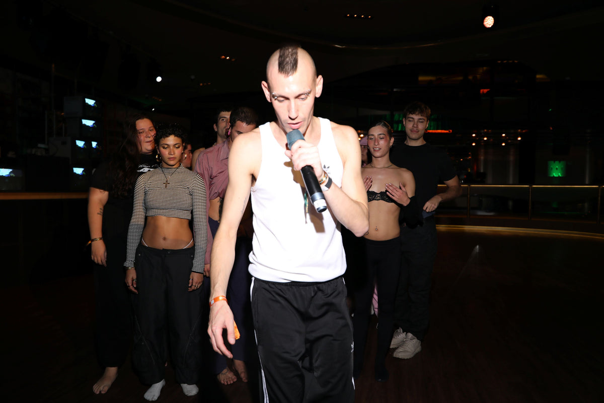 A young man is holding a microphone in his hand and singing. In the background you see a group of people all looking in the direction of the artist.