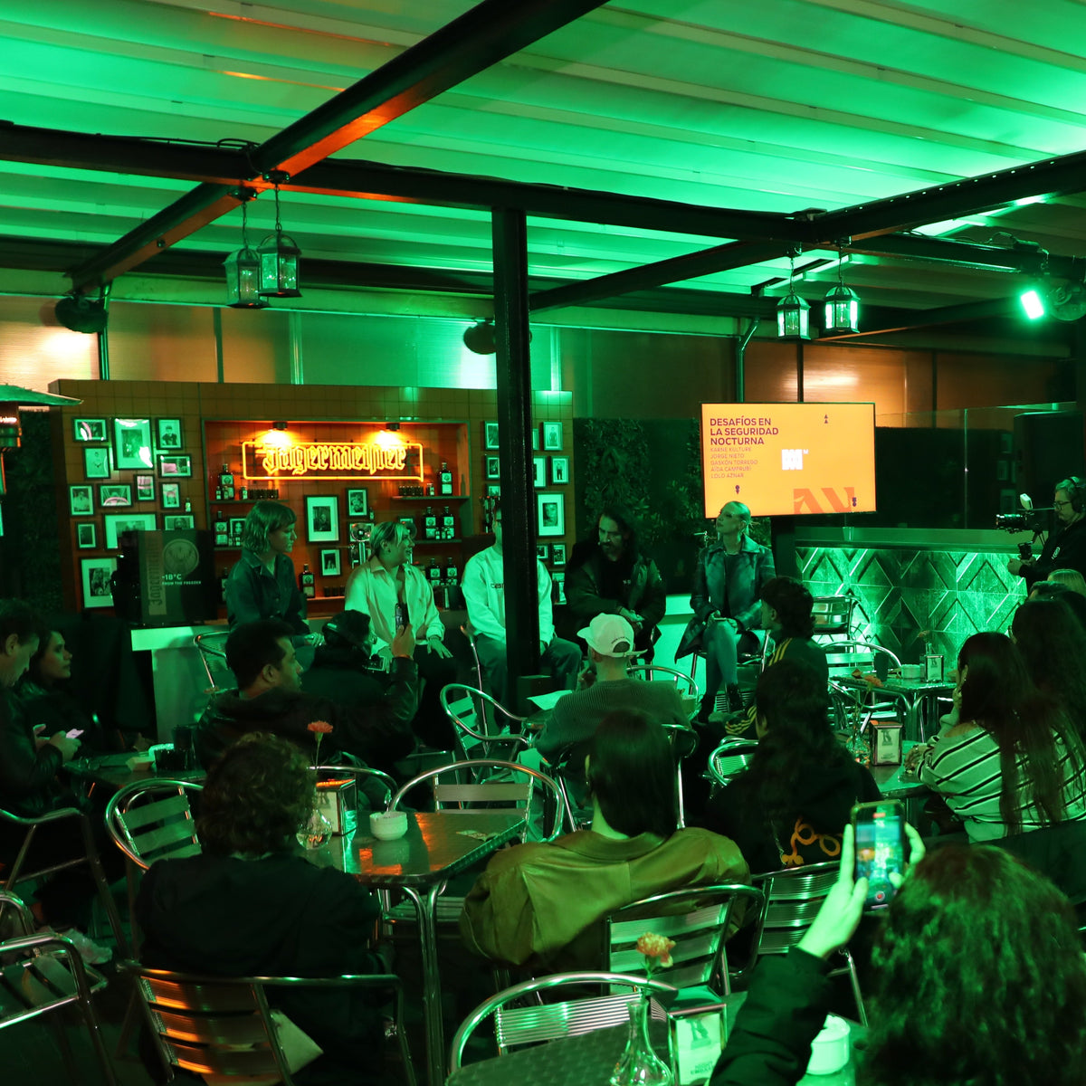 A crowded indoor event or lecture hall scene with many seated and standing people facing a stage area. The room is heavily lit with green ambient lighting, and a screen is visible on the stage.