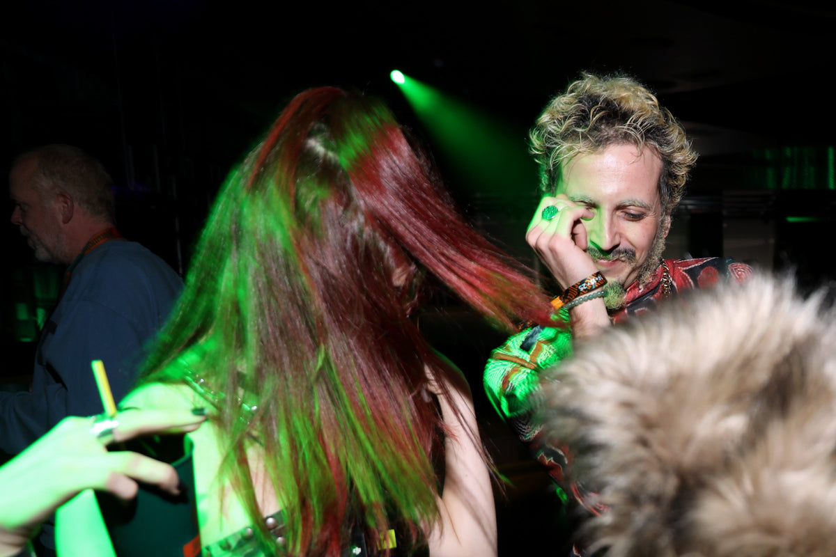 A person with long red hair stands in a dark nightclub or event space under green stage lighting, facing a man with curly blond hair and a mustache who is touching his face. Another person is partially visible on the left, and a furry foreground object intrudes at the bottom right.