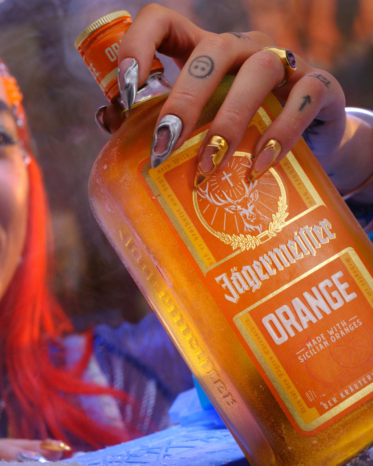 A close-up lifestyle shot of a person holding a bottle of Jägermeister Orange liqueur. The person has bright red hair, a nose ring, and silver and gold manicured nails.