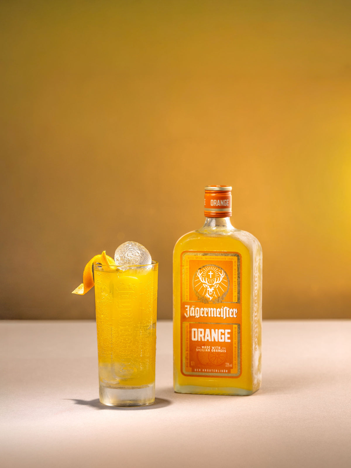 A bottle of Jögermeister Orange liqueur stands next to a highball glass filled with the orange-colored drink, garnished with an orange peel twist and a large ice sphere, set against a warm yellow gradient background.
