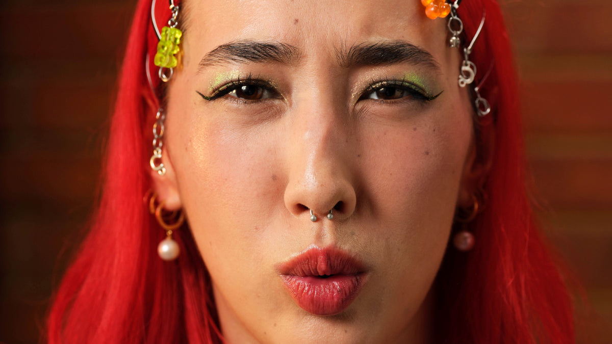 Close-up portrait of a person with bright red hair, green eye makeup, a nose ring, and colorful dangling earrings against a warm brown background.