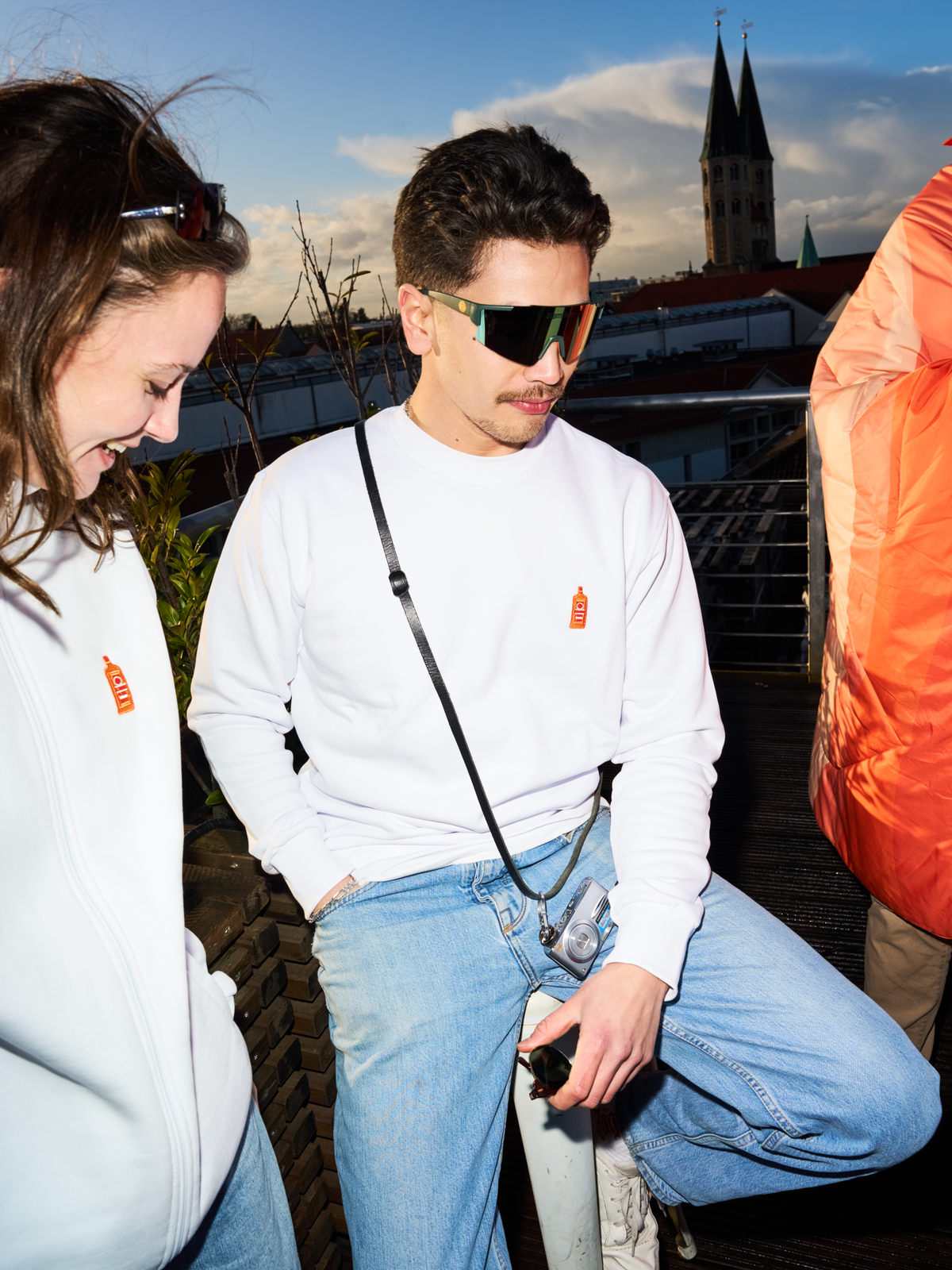 Two People wearing white Jägermeister sweatshirts and Jägermeister fast glasses on a rooftop terrace, with a church tower and city skyline in the background.