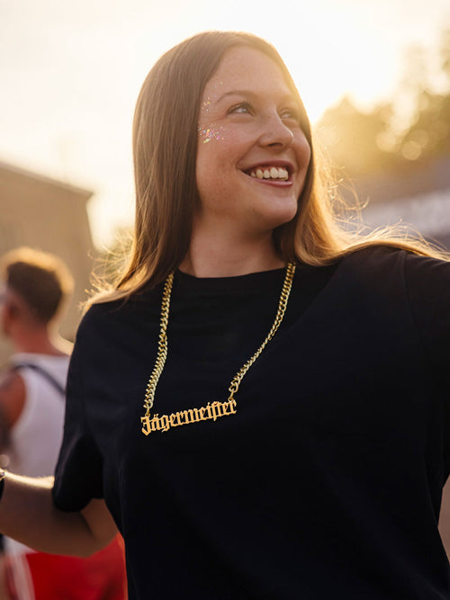 Woman wearing a black shirt and a gold chain necklace with a Jägermeister charm, smiling outdoors in a festival setting.