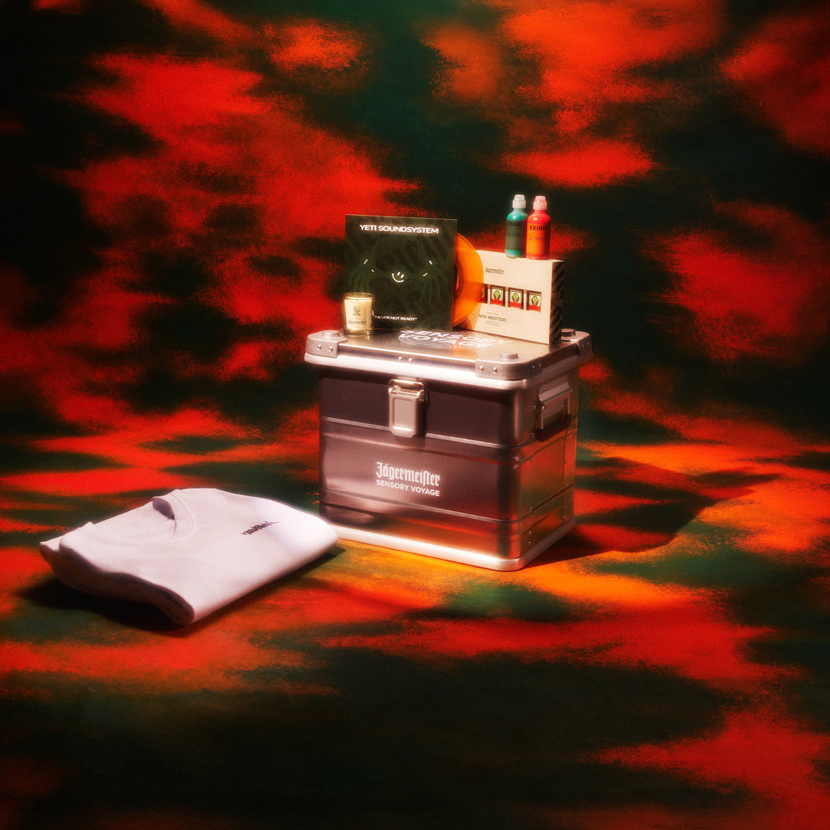 A dark metal storage trunk sits on a red and green mottled surface with a white folded shirt beside it and small items on top, including a black box labeled 'Jägermeister - Sensory Voyage'.