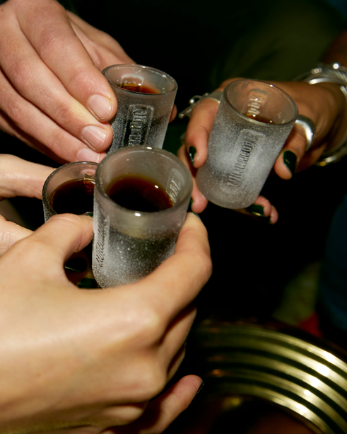 Four hands holding frosted Jägermeister shot glasses filled with a dark liquid, clinking together.