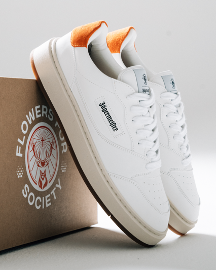 A pair of white low-top sneakers with orange heel tabs and black 'Jägermeister' branding on the side, displayed next to a brown cardboard shoebox printed with 'FLOWERS FOR SOCIETY'.