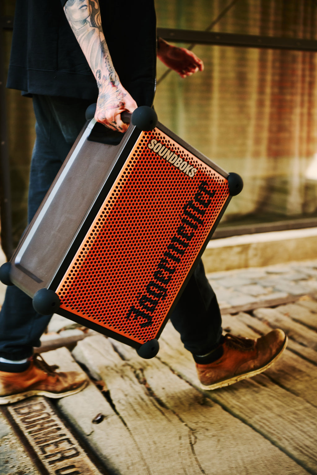 Person holding an orange and black portable Soundboks speaker with 'Jägermeister' branding walking on a wooden floor