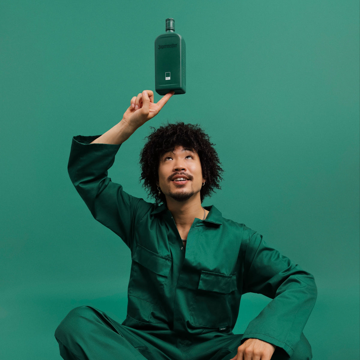 A man in a dark green jumpsuit balances the Jägermeister herbal green pantone bottle on his fingertips against a solid teal-green studio background.