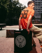 A man in an orange Jägermeister jersey and beige pants sits on a black Soundboks speaker with Jägermeister brand logo