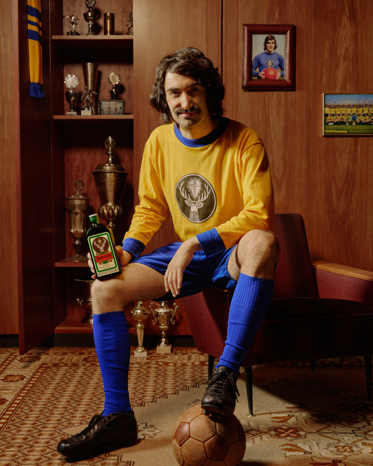 A man wearing a vintage-style yellow and blue soccer jersey, blue shorts, and blue socks sits in a wooden room holding a Jägermeister retro-bottle. The jersey features a circular deer logo on the chest.
