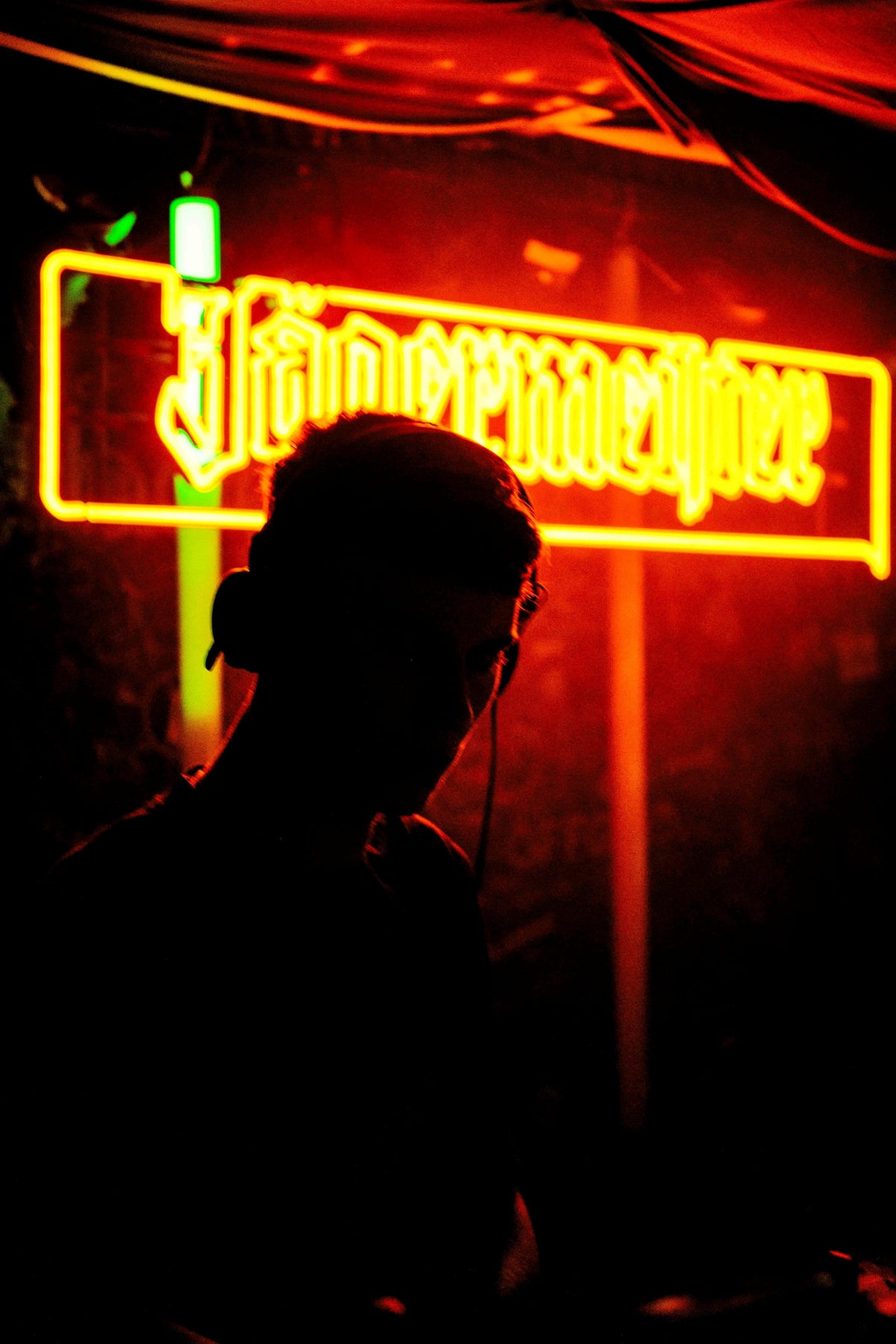 Silhouette of a DJ standing in front of a glowing Jägermeister neon sign in a dimly lit club.