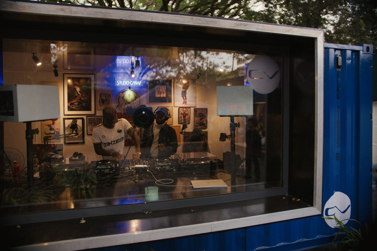 A blue shipping container kiosk with a large front window, showing a DJ setup inside with two people operating turntables. The container has a black frame and white circular logo decals on the side.