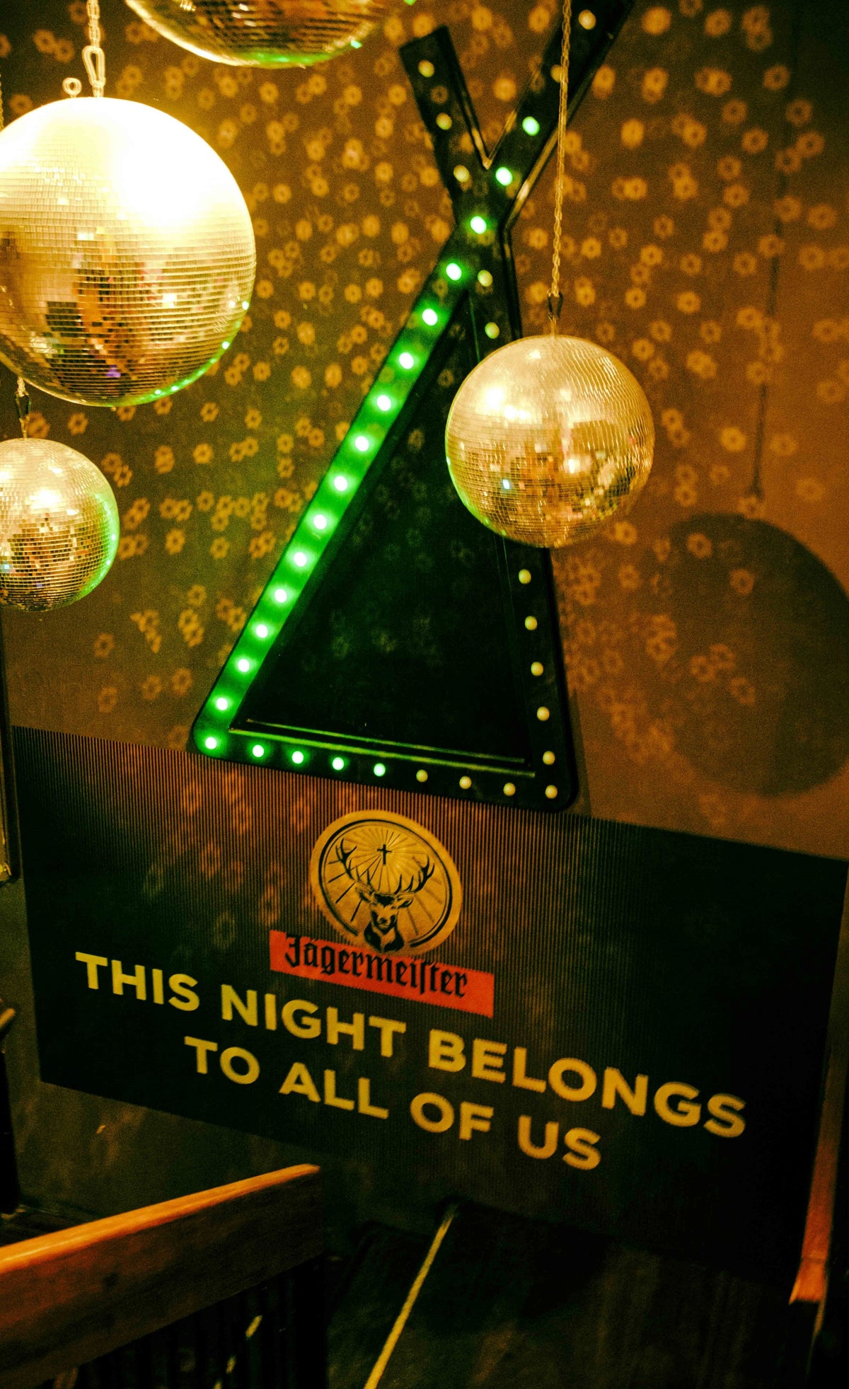 A Jägermeister sign reading 'THIS NIGHT BELONGS TO ALL OF US' with a green illuminated triangle outline, surrounded by hanging disco balls against a dark patterned background.